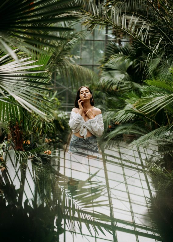 A woman stands gracefully amidst tropical plants in a greenhouse, evoking tranquility.